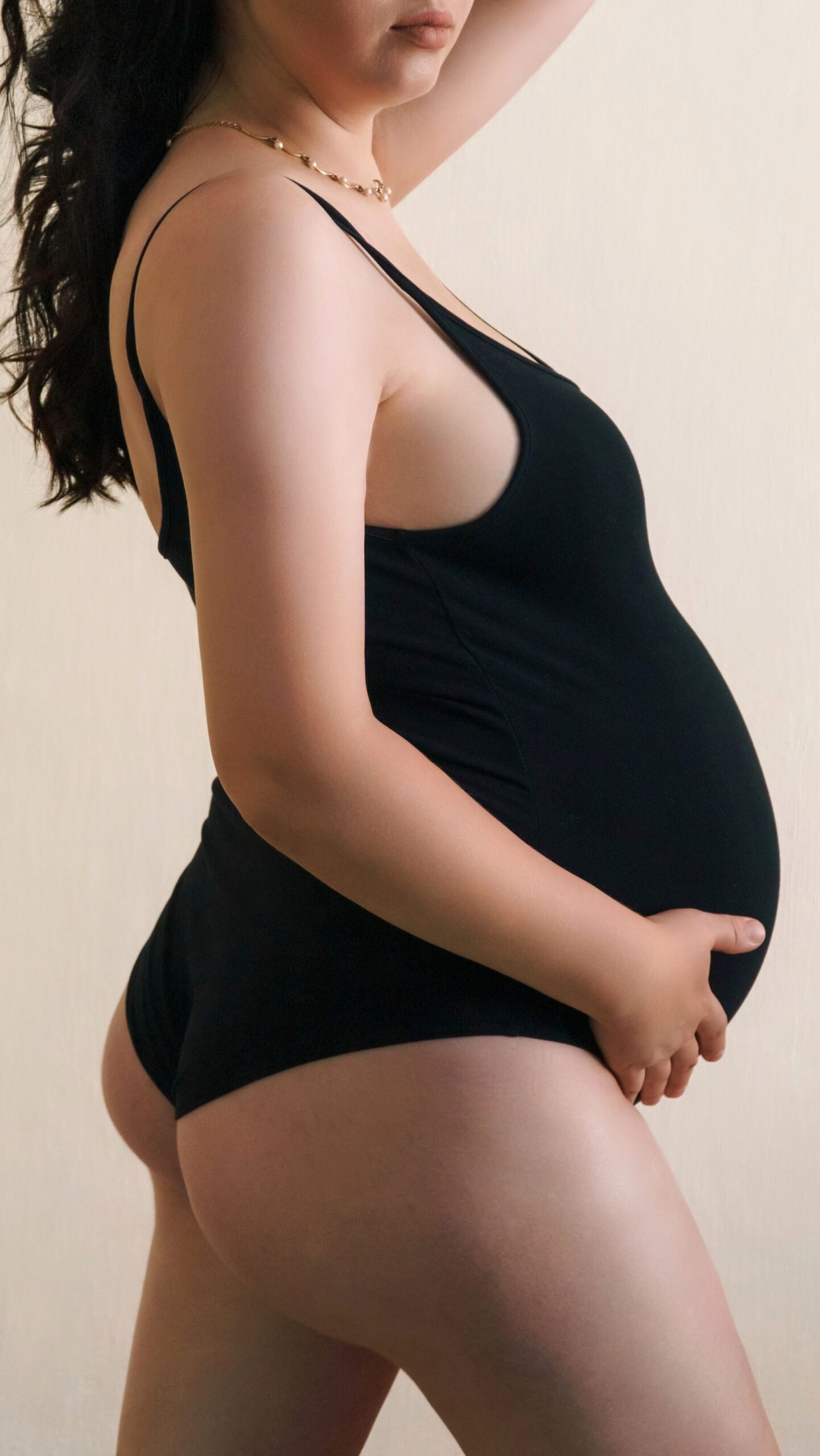 Portrait of a pregnant woman wearing a black bodysuit posing confidently against a neutral background.